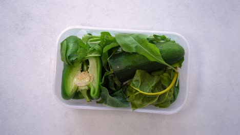 Overhead-View-Of-Hand-Putting-The-Tub-Of-Green-Vegetables-On-White-Table