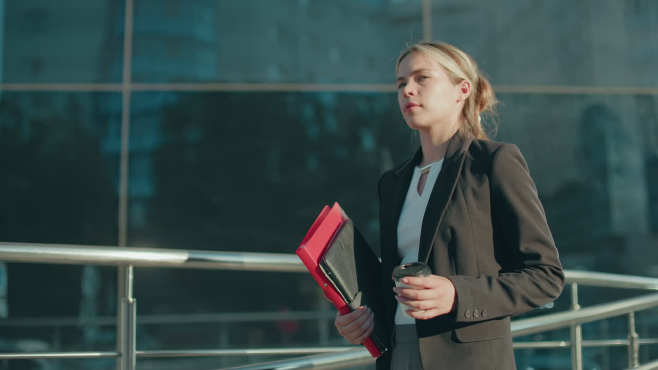 Side view of young lady in business attire walking while holding file folder and coffee cup with bright sunlight ambiance and reflective metal railing in front of modern glass building background