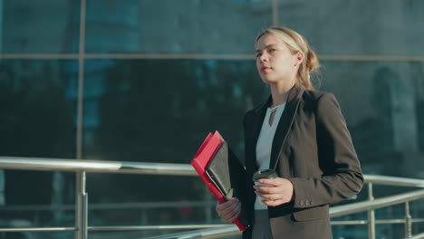 side view of young lady in business attire walking while holding file folder and coffee cup with bright sunlight ambiance and reflective metal railing in front of modern glass building background