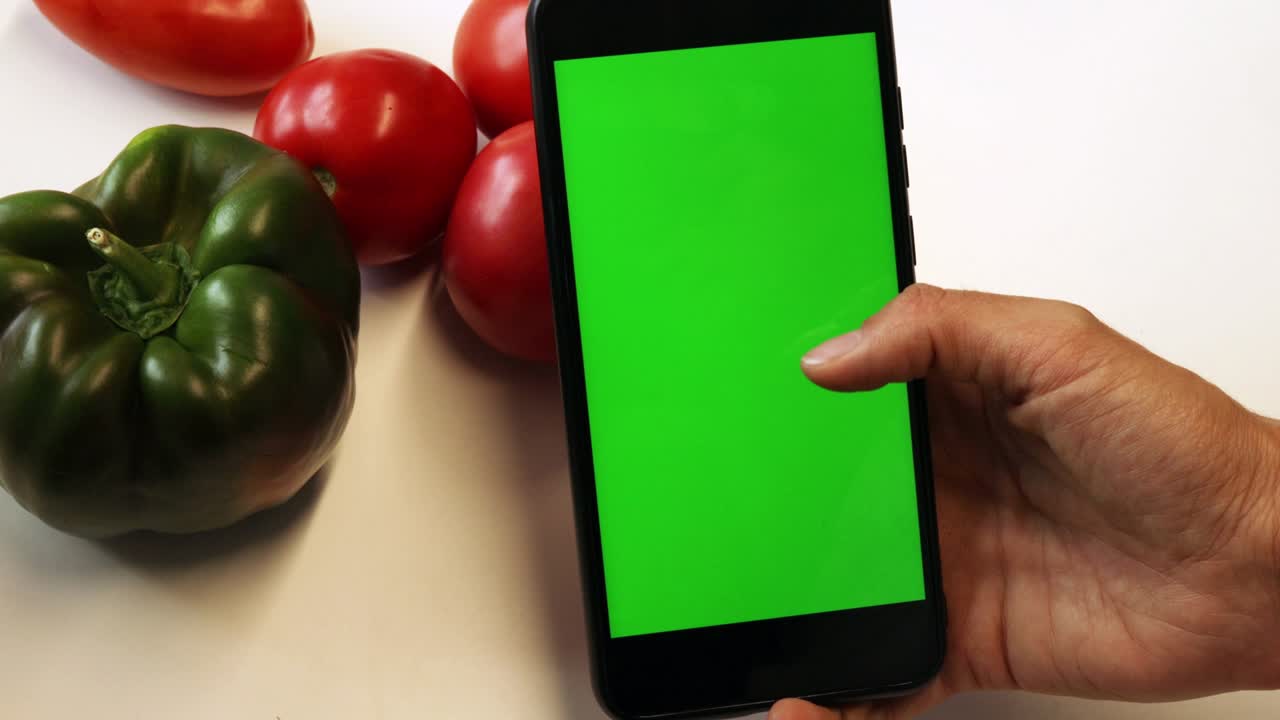 primer plano de la mano de una mujer sosteniendo una maqueta verde de pantalla de teléfono inteligente. teléfono móvil moderno. comida orgánica en el fondo. verduras, tomate y pimienta. espacio de copia. llave de croma