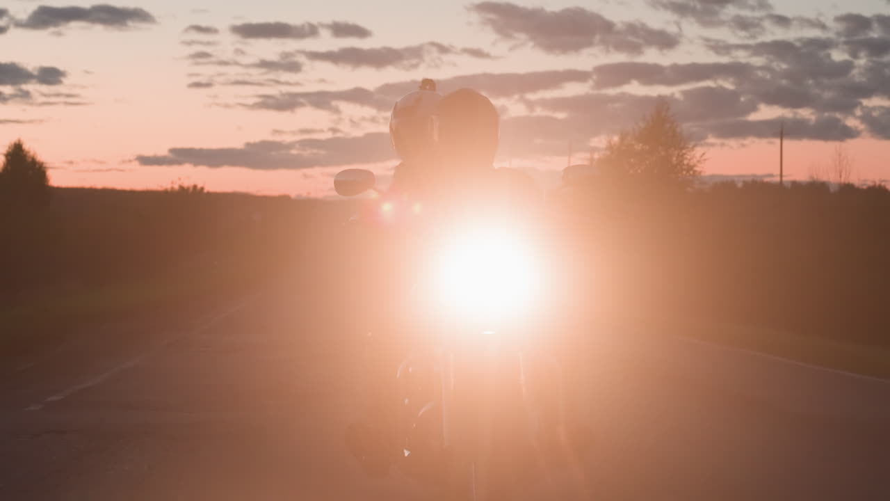 Motorcyclist carrying passenger riding countryside road at evening sunset with bright headlamp glowing ahead, silhouette visible against dramatic sky as journey continues through open landscape
