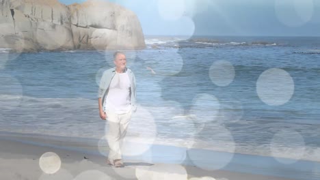 Animation-of-glowing-lights-over-a-sad-senior-caucasian-man-walking-on-the-beach