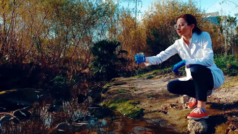 a young woman scientist at a creek, wearing protective eyewear and a lab coat, taking a water sample from the creek with a beaker