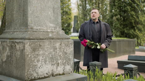 man in black raincoat and suit holding red roses standing in front of a tombstone in a graveyard 2