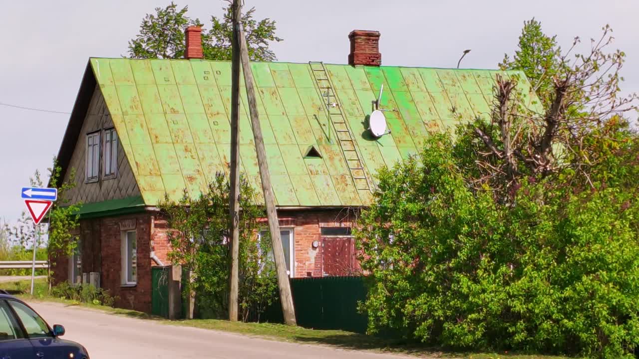 Old red-brick house with a green corrugated metal roof, satellite dish, and traffic sign on a village road in rural Latvia. Location: Latvia (Latvija)