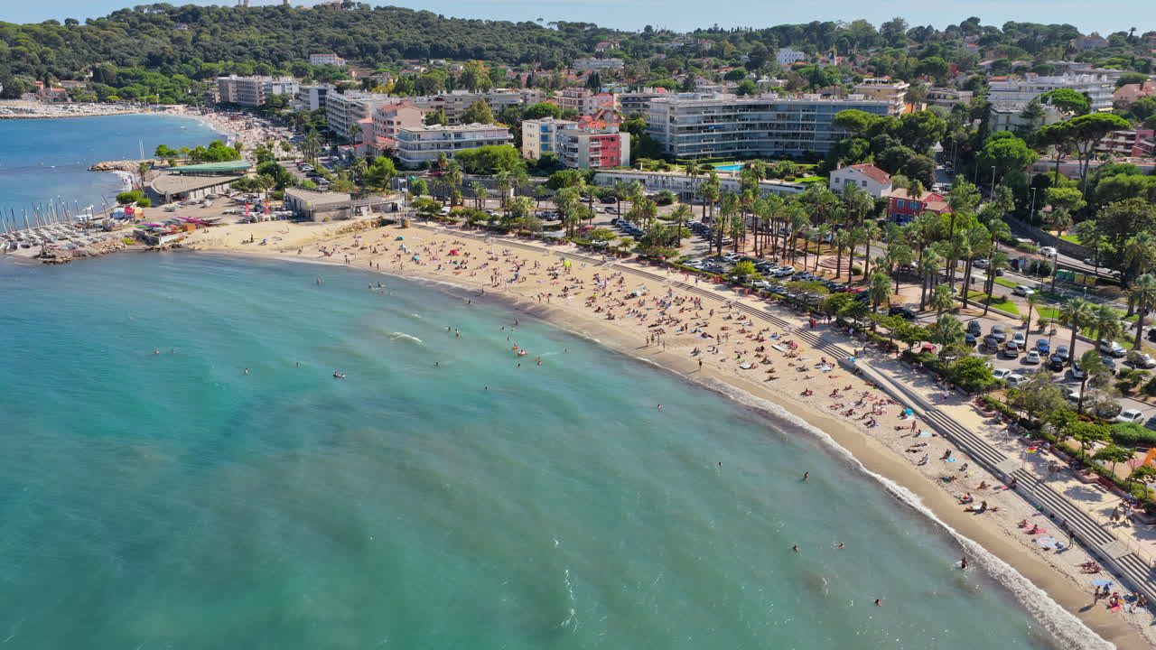 Vista aerea da drone della spiaggia e del lungomare di Antibes, fiancheggiati da palme e appartamenti, con acque turchesi del Mediterraneo e bagnanti nella baia