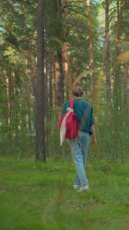 dama camina lentamente a través de sereno bosque iluminado por el sol, mochila roja colgada sobre su hombro, con camisa verde, ella parece cansada, mirando por encima del humbro, como la luz del sol filtra a través de los árboles y la hierba alta