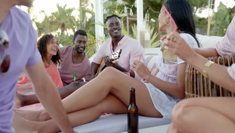 Happy-diverse-group-of-friends-relaxing-with-guitar-and-drinking-cocktails-and-beers-at-beach