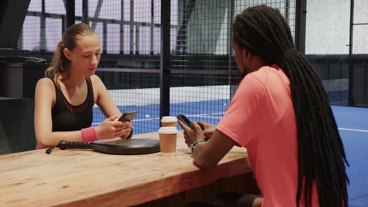 Due atleti diversi discutono la strategia del padel tennis davanti a un caffè al campo indoor.