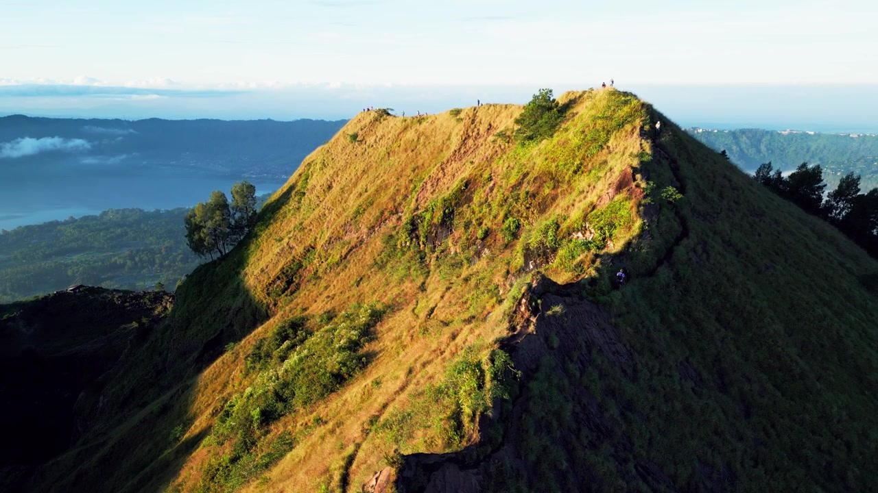 Jalur gunung dengan pendaki matahari terbit melintasi medan vulkanik curam di atas kaldera Gunung Batur yang diselimuti kabut dan dataran tinggi Bali yang dramatis, pengambilan gambar drone udara dengan gerakan maju.
