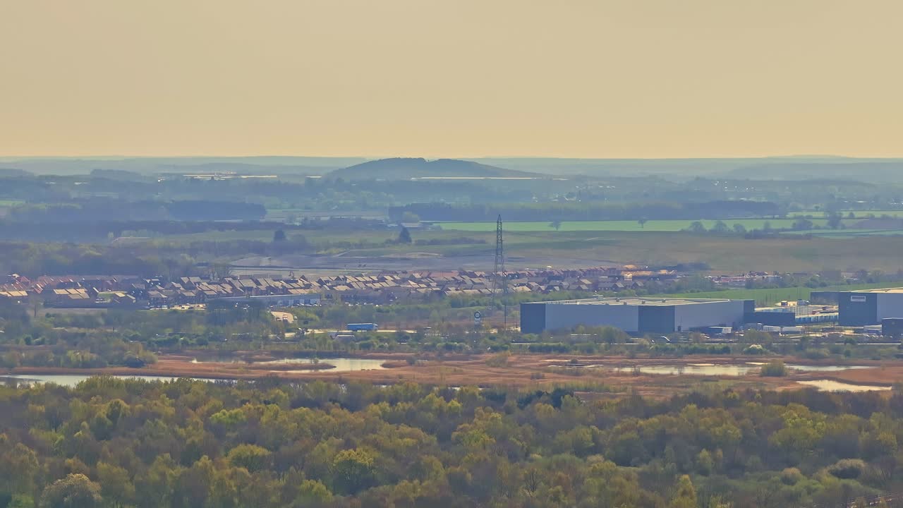 Drone ascends showing dense woodland, Doncaster suburban outskirts, industrial buildings, and distant rolling hills under soft golden sky in evening