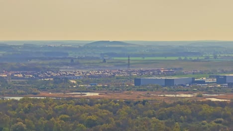 drone ascends showing dense woodland, doncaster suburban outskirts, industrial buildings, and distant rolling hills under soft golden sky in evening