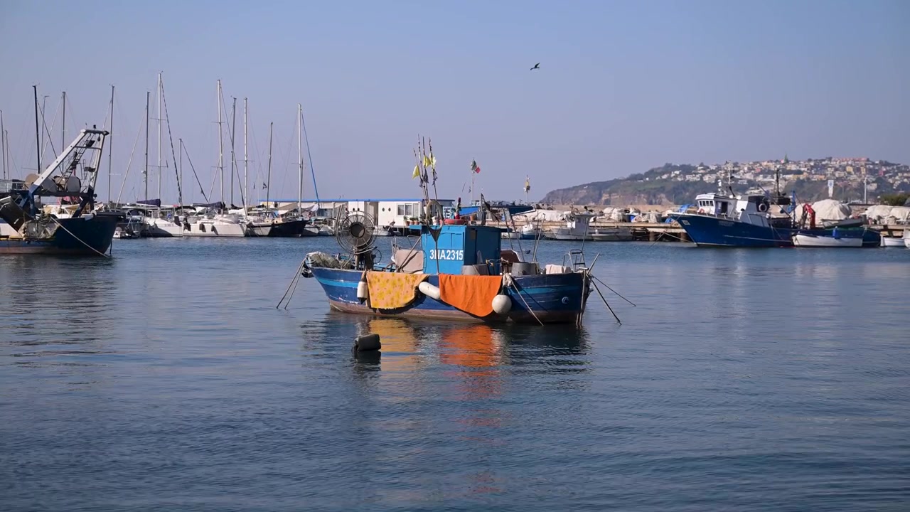 Una piccola barca da pesca blu con reti colorate galleggia in un tranquillo porto turistico italiano con yacht e case sulla collina sullo sfondo