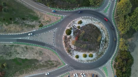 Aerial-view-of-a-roundabout-with-several-vehicles-of-different-colors-turning,-highlighting-traffic-organization,-road-infrastructure,-and-various-colored-vegetation