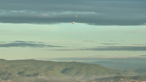 Far-away-view-of-colorful-hot-air-balloons-flying-over-mountain