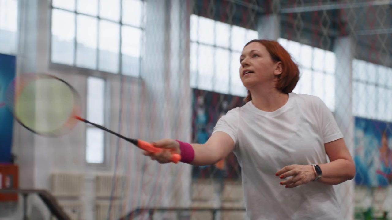 Indoor badminton court practice with woman holding racket near net, pink wristband, white shirt, red hair, calm focused warmup, gentle serves and short rallies, light breathing, community sports hall