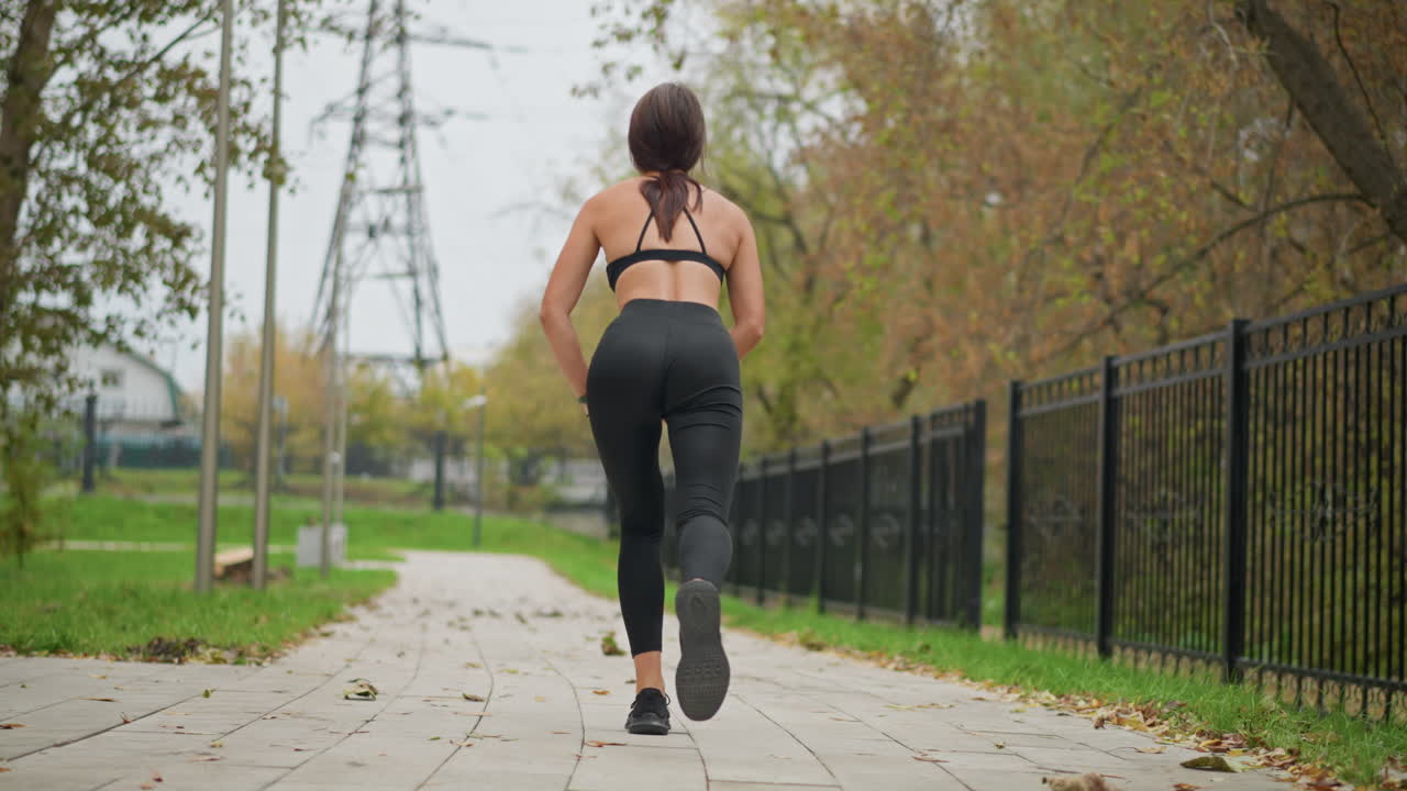 Back view of woman stretching her leg outdoors in park wearing black leggings and a sports top, she is exercising in a park pathway surrounded by trees, iron fence, and a peaceful environment