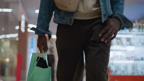 front shot of young man walking through modern shopping mall carrying shopping bag wearing denim jacket hoodie crossbody bag focus on hand and torso bokeh background urban shopping lifestyle