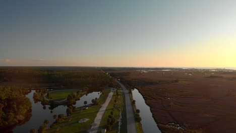 Vista-Aérea-De-Un-Largo-Camino-Construido-Entre-Dos-Lagos,-Con-Algunos-Autos-Conduciendo-Hacia-Un-Gran-Bosque,-Disparo-Al-Atardecer-En-Florida