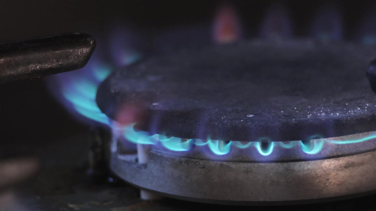Una perspectiva lateral de primer plano extremo de un quemador de gas de cocina encendiéndose en un anillo de llamas azules constantes. Toma macro con trípode fijo. Ambiente interior