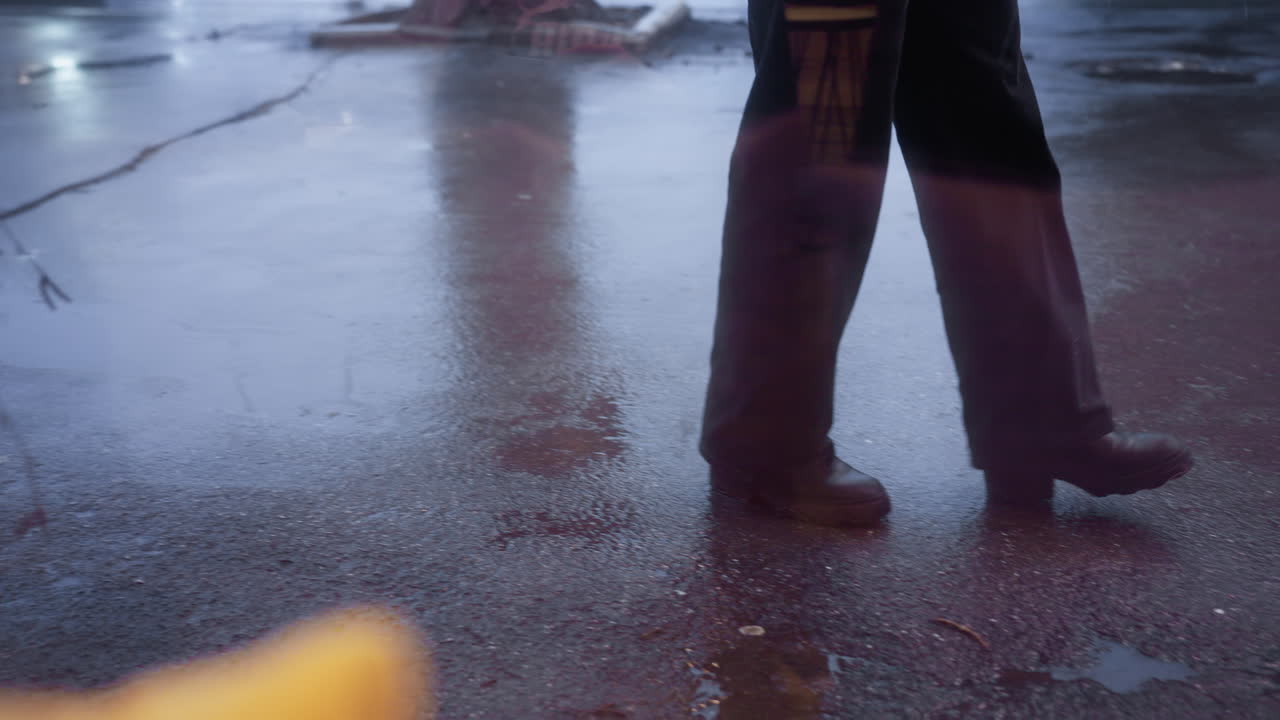 Close-up of person in black trousers and boots walking calmly on wet ground during rainy weather, with a focus on legs, reflecting the rainy mood and wet pavement around them