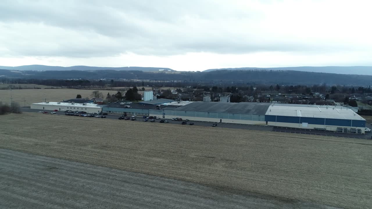 A backward moving drone shot capturing commercial buildings beside in front of a large green field, with houses visible across the background of the scene