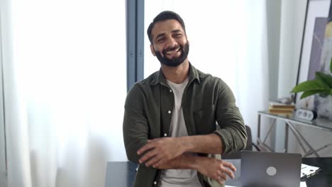 retrato de un hombre carismático, positivo, exitoso, indio o árabe, director ejecutivo de una empresa, freelance o diseñador creativo, de pie en su oficina moderna con los brazos cruzados, mirando a la cámara, sonriendo amistosamente