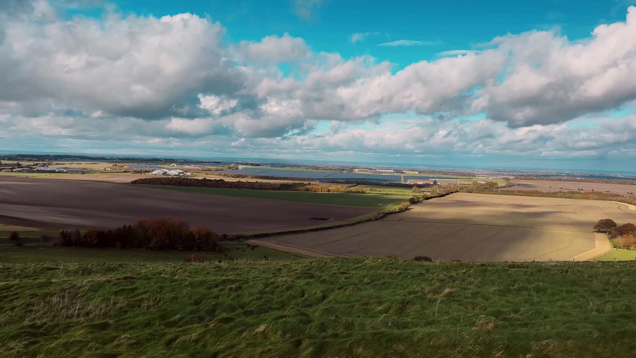 Drone video capturing panoramic view of English patchwork farmland and water. Wide-angle shot from elevated grassy hill under vibrant blue skies with scattered clouds