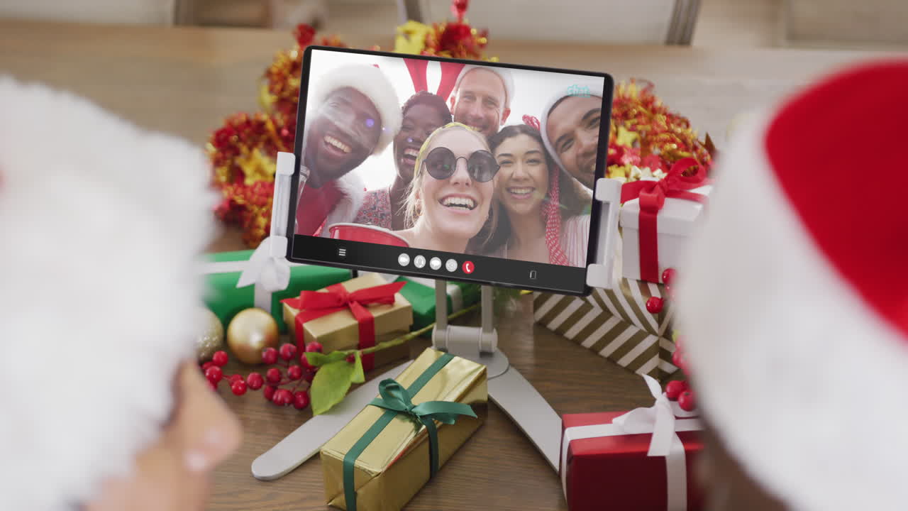 animación de una pareja diversa con sombreros de papá noel en una llamada de video de tableta con amigos en navidad