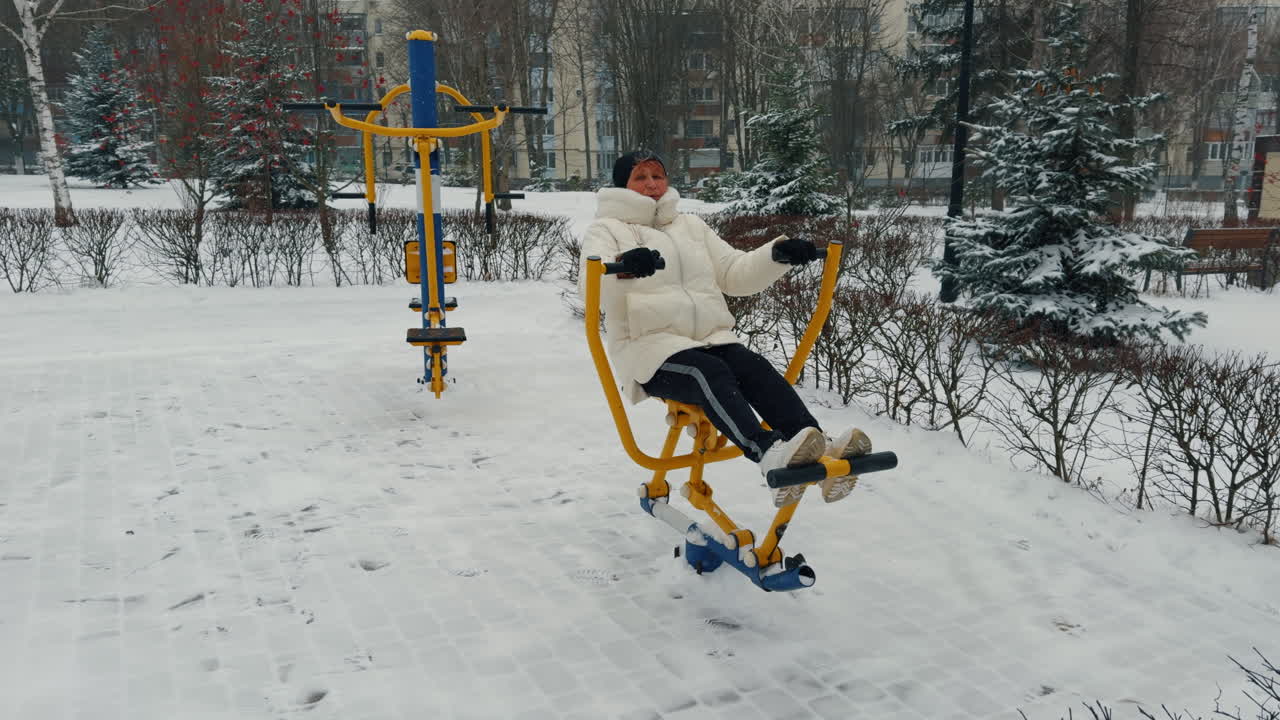 Caucasian old woman exercising on the simulator performing rowing movements. Winter training outdoors.