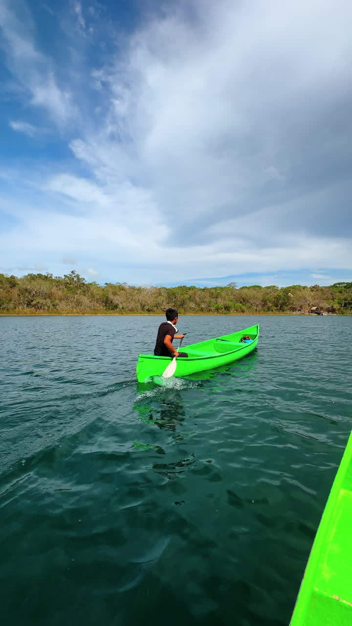 Man paddling green canoe on calm lake under cloudy sky. Rear view of a man rowing a bright green boat on a wide body of water with forest horizon