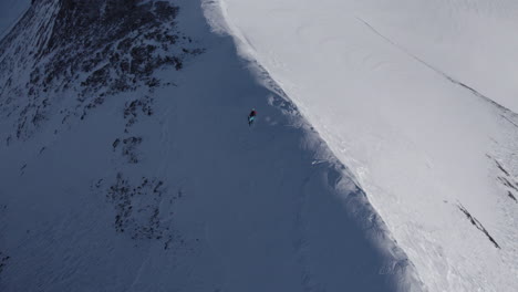 Toma-Aérea-De-Arriba-Hacia-Abajo-De-Un-Esquiador-Solitario-Tallando-Una-Montaña-Nevada-Y-Rocosa-Extremadamente-Empinada-Durante-El-Sol