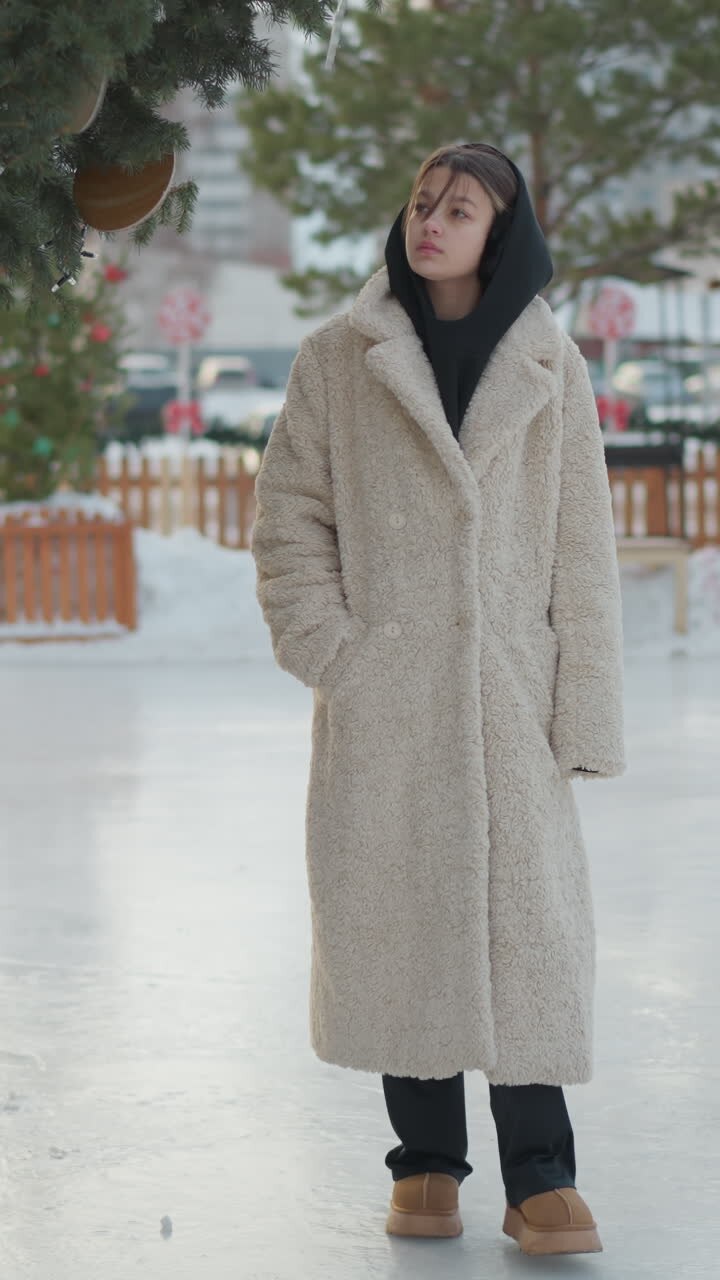Giovane donna con cappotto shearling in piedi su una piazza ghiacciata vicino a un sempreverde decorato, che guarda in su con le mani in tasca, luce soffusa e nuvolosa, mercatino natalizio urbano sullo sfondo, atmosfera serena e contemplativa