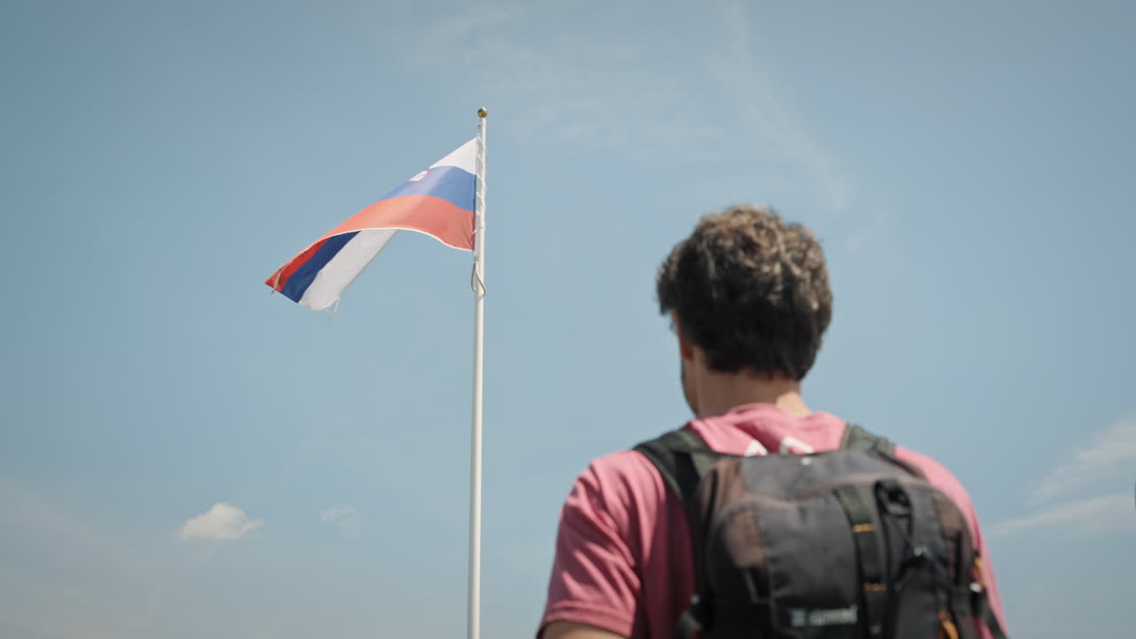 пеший турист из йонга заслоняет взгляд от солнца и смотрит на шест с флагом словении, который развевается на ветру