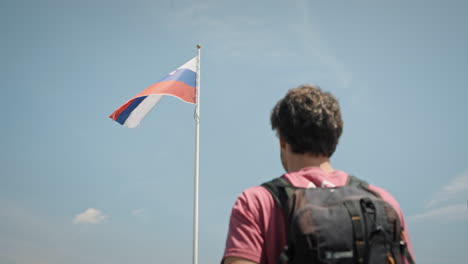 a yong hiker shading his glance from the sun and looking at the pole with slovenian flag which is fluttering in the wind