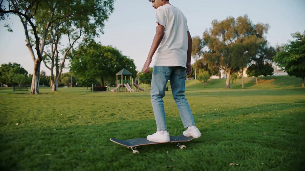 Placing skateboard teen in jeans stepping on deck rolling slowly across park grass, copy space