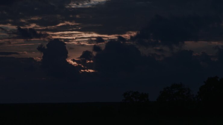 Ripresa in time-lapse di un cielo crepuscolare drammatico mentre il sole tramonta dietro nuvole fitte, con raggi di luce che filtrano e alberi in silhouette lungo l'orizzonte