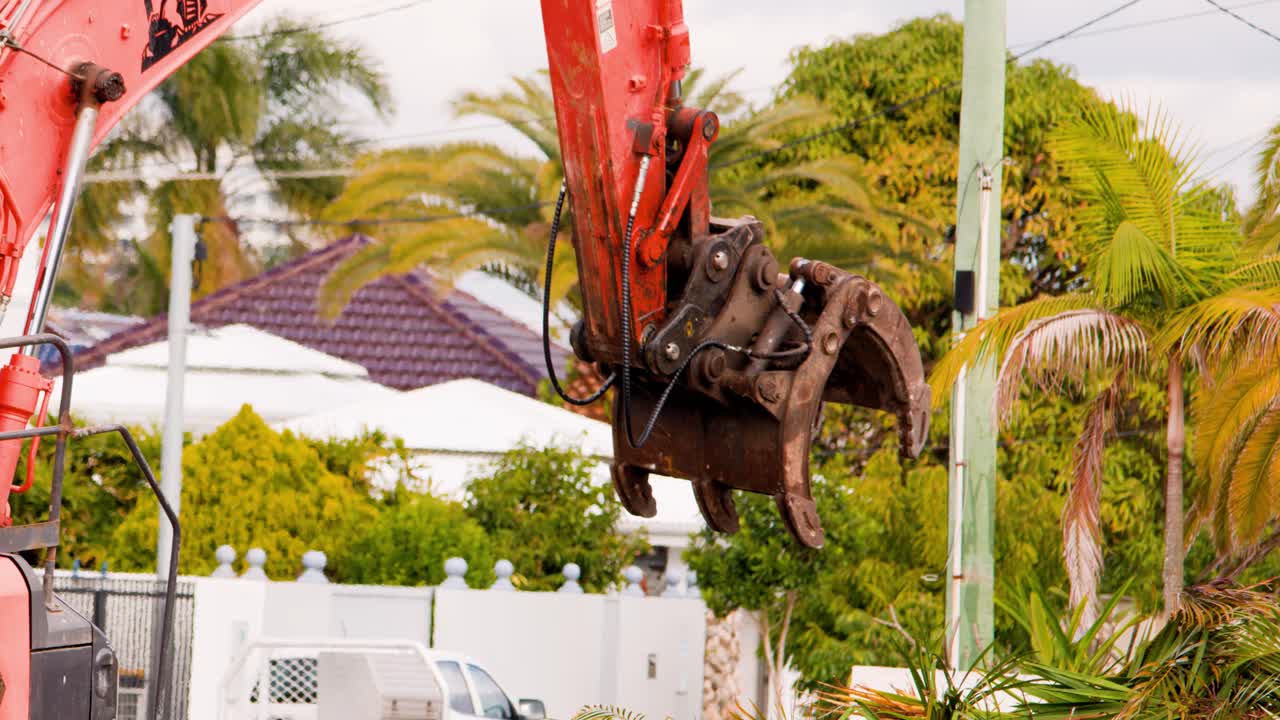 An orange excavator uses its hydraulic claw to remove a palm plant from the ground at a suburban construction site on a cloudy day