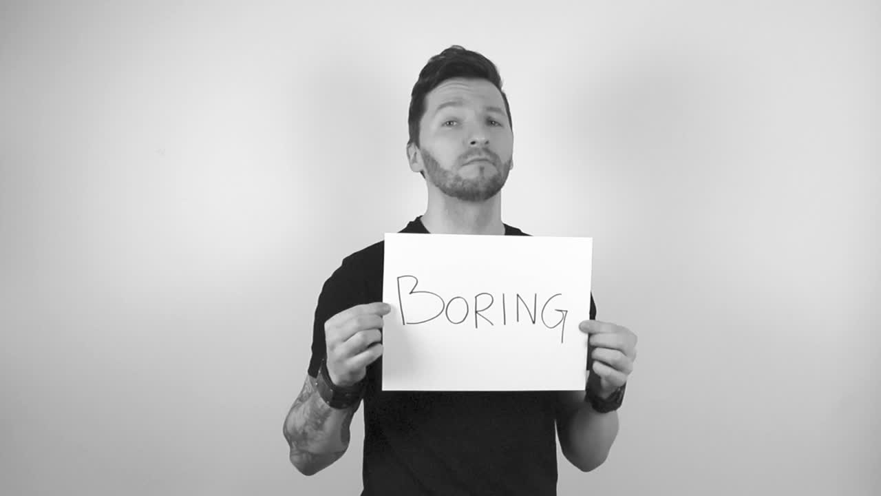 Grayscale footage of a tattooed man with short hair and beard lifting up a white paper or a sign that says BORING on it in capital letter. Man is seemingly bored about something, while looking forward