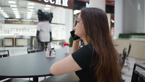 side view of a woman on a phone call, resting her hand on a table in a busy mall, with an unfocused background enhancing the focus on her