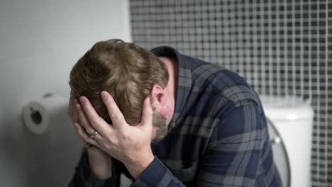 constipated man sits on toilet with his head in hands, suffering in pain