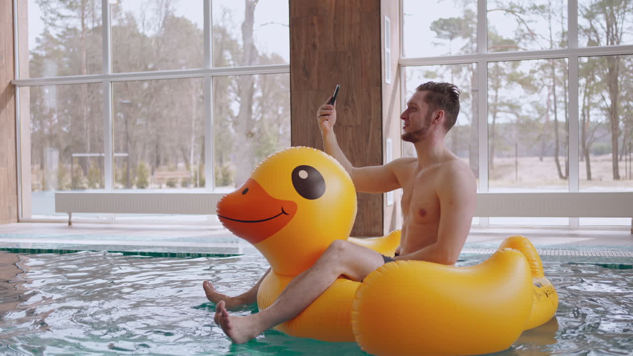 hombre feliz está sentado en un pato inflable divertido en la piscina y hablando por videollamada en un teléfono inteligente