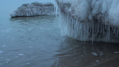 Small-waves-breaking-against-the-ruins-of-Karosta-Northern-Forts-fortification-on-the-shore-of-Baltic-sea-on-a-cloudy-winter-day,-covered-with-ice,-snow-and-icicles,-closeup-shot