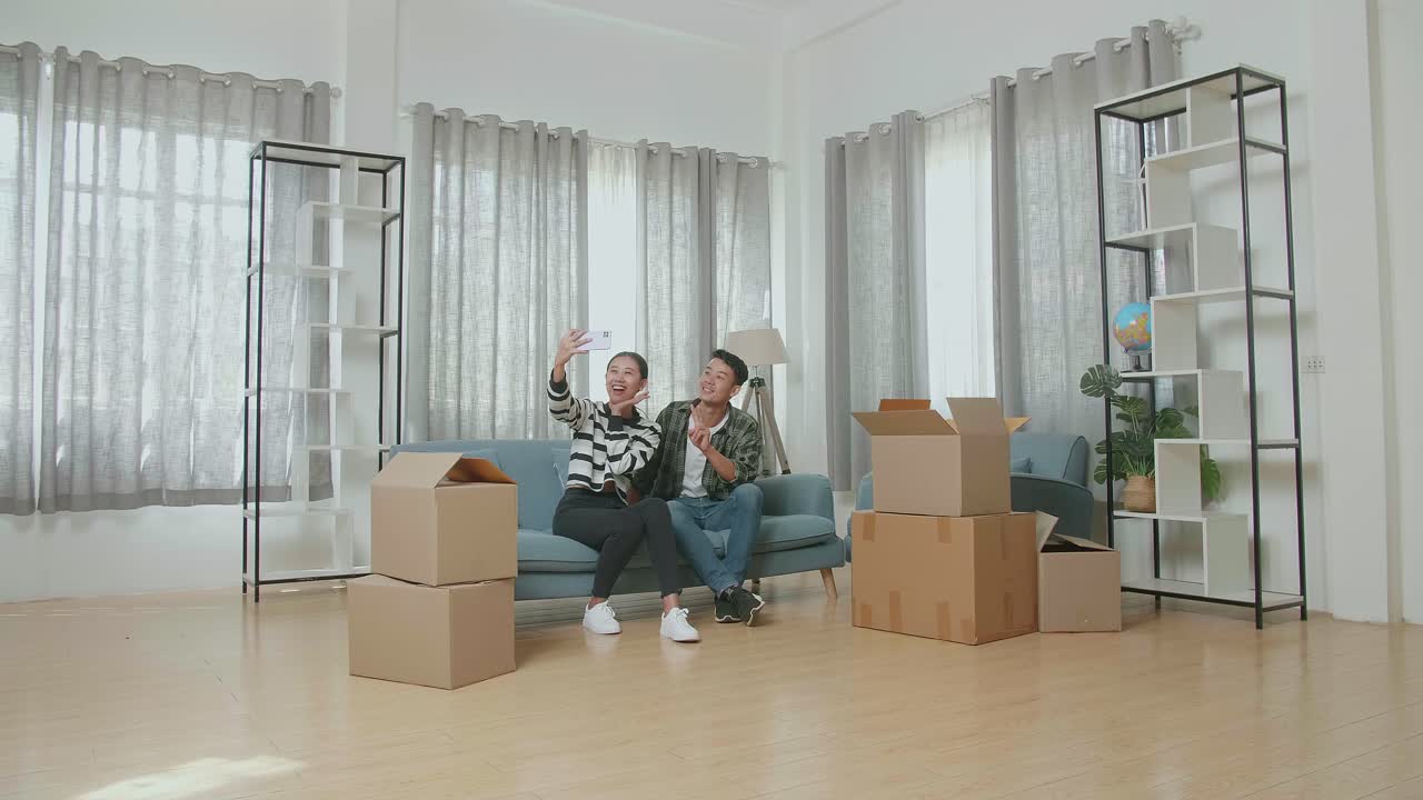 una joven pareja asiática con cajas de cartón en el suelo sentada en un sofá y tomando fotos con su teléfono inteligente después de mudarse a una nueva casa