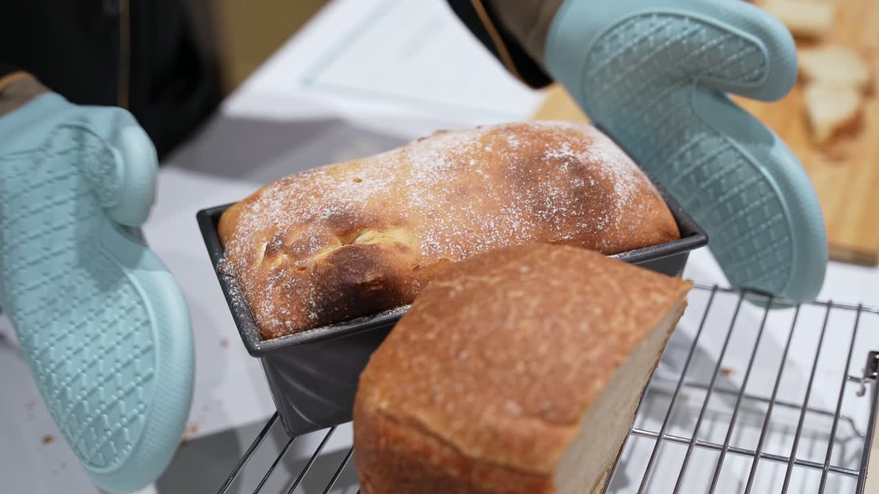 Close-up of an oven-mitt hand probing a baked loaf with a thermometer in a bread pan on the counter, indoor kitchen scene under bright light