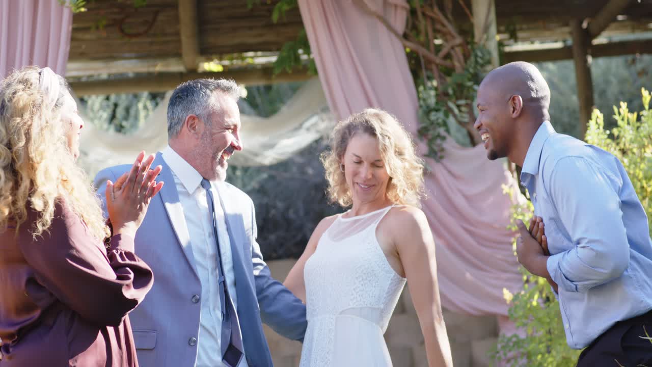 Una coppia in abiti eleganti con ospiti eterogenei dopo gli applausi, sotto un pergolato, con la sposa che volteggia e lo sposo che ride.