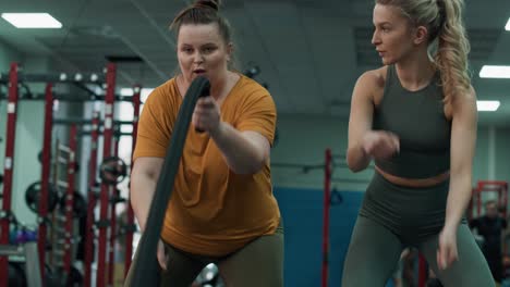 mujer con sobrepeso ejercitándose con cuerdas de batalla y entrenadora en el gimnasio.