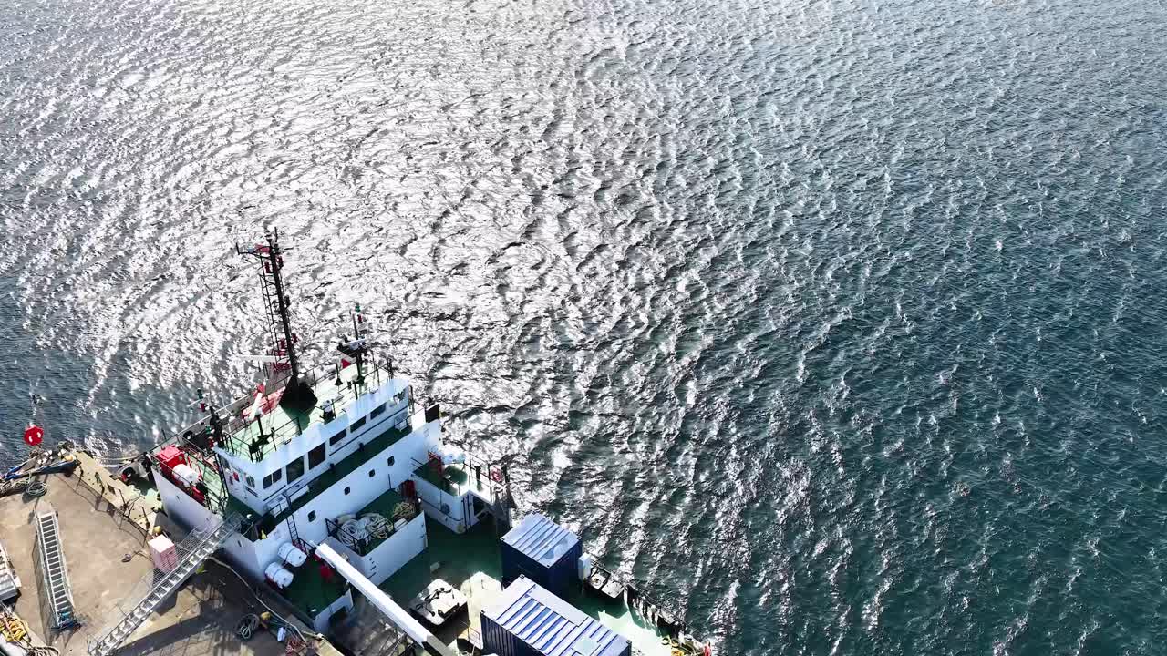 Drone ascends over maritime pier, revealing fishing vessel, industrial dock, and shimmering coastal waters