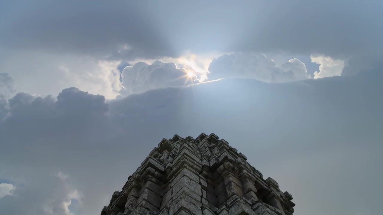 雲の隙間から光線が差し込み、寺院に立つ彫刻された石の塔が日光を浴びている。