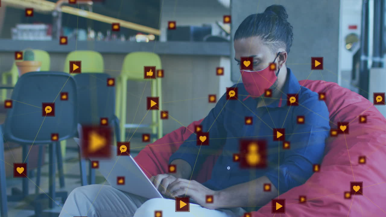 animación de una red de iconos digitales sobre un hombre biracial usando una máscara facial usando una computadora portátil en la oficina.